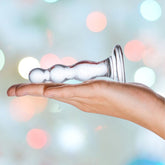 A hand holds the transparent Gläs Triple Play 4" Beaded Glass Butt Plug Dildo with a flared base, set against a backdrop of blurred, colorful lights.